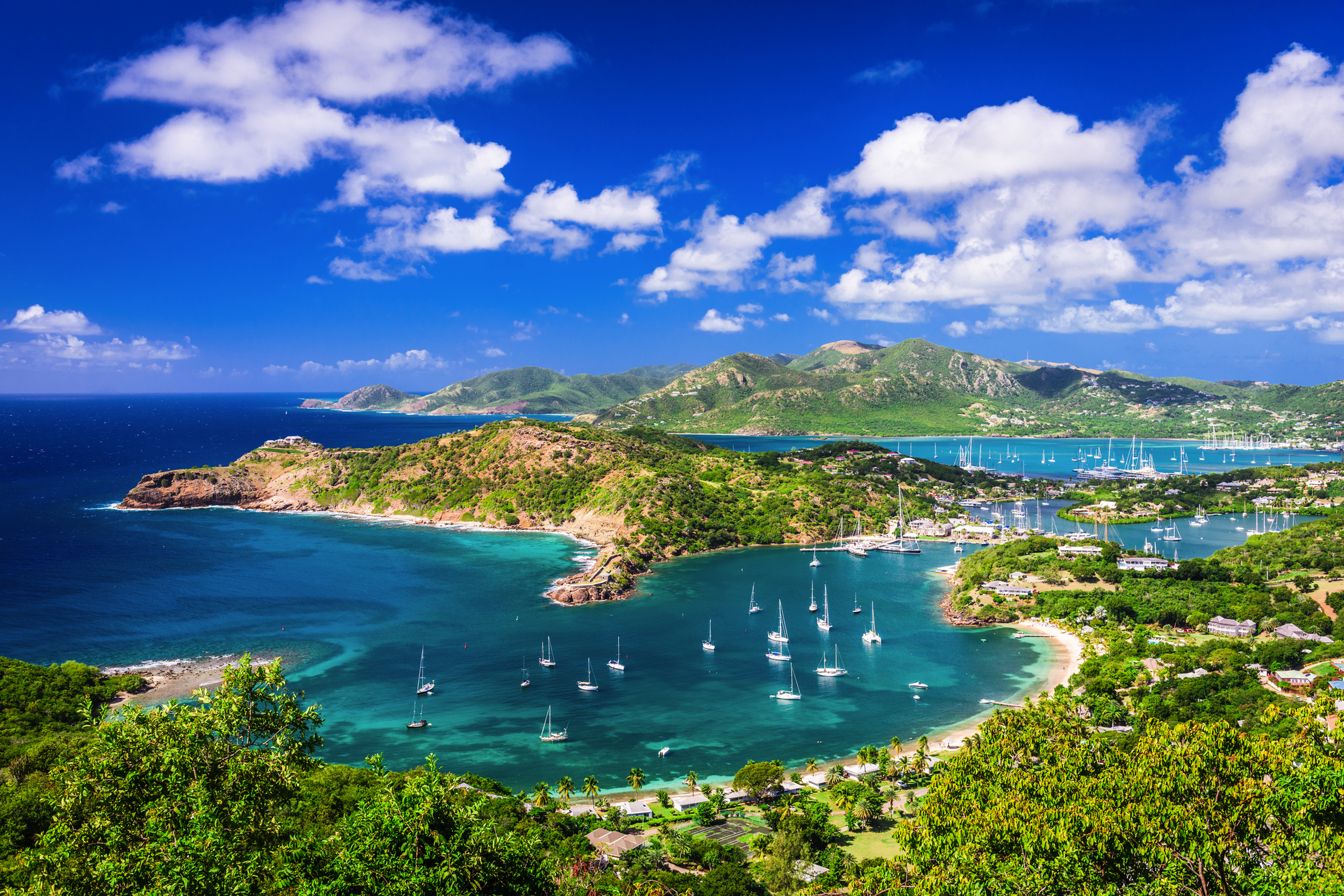 Küstenlandschaft mit türkisem Wasser und zahlreichen Segelbooten, umgeben von grünen Hügeln.
