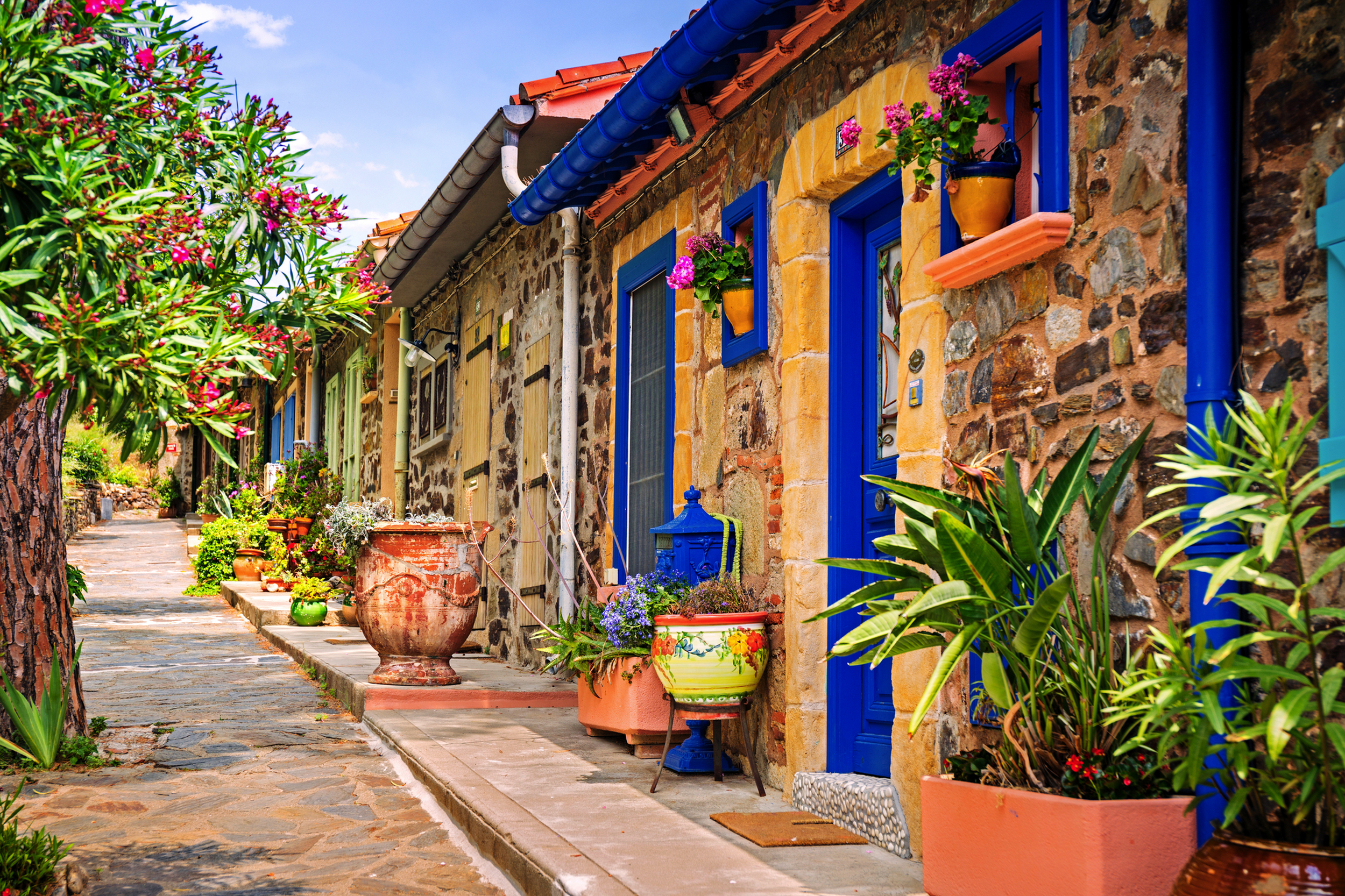 Bunte Häuserfassaden mit Blumen vor steiniger Wand entlang eines gepflasterten Weges.