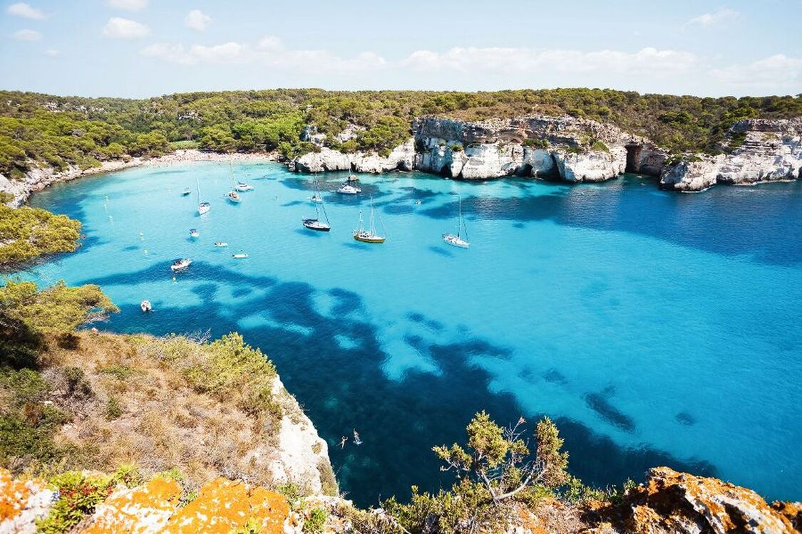 Küste mit türkisfarbenem Wasser, Booten und bewaldeten Klippen im Hintergrund.