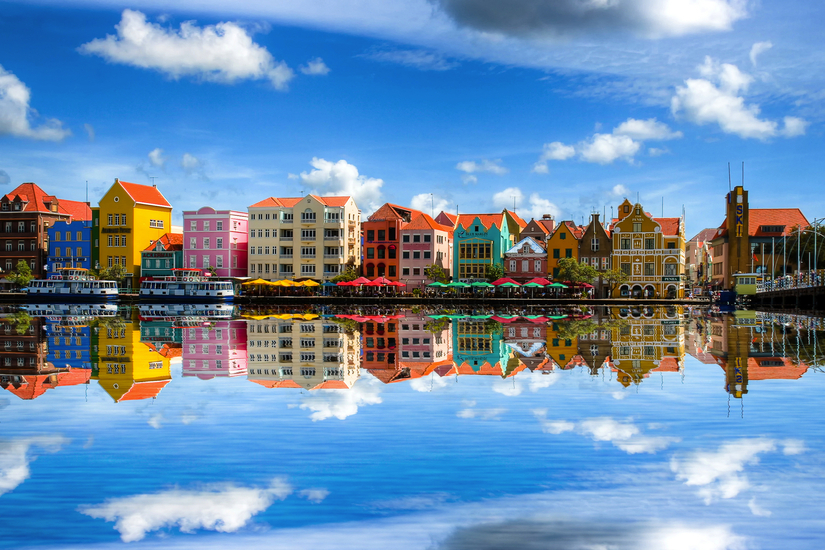 Farbige Häuserzeile spiegelt sich in Wasser unter blauem Himmel mit Wolken.